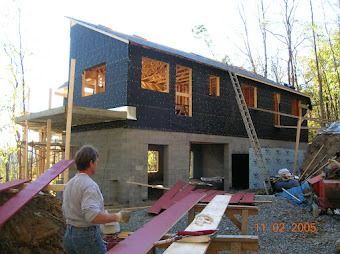 House under construction with dark siding, ladders, and a man carrying long boards outdoors