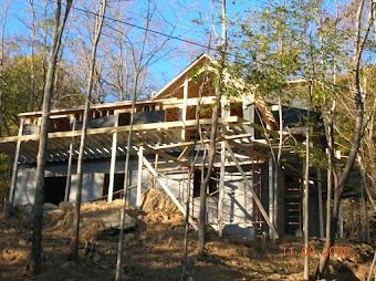 House under construction in a wooded area, with exposed framing and a roofed upper level.