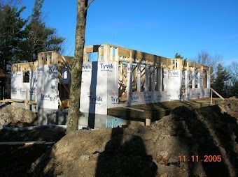 Partially framed house under construction with Tyvek wrap and exposed wooden beams on a dirt lot