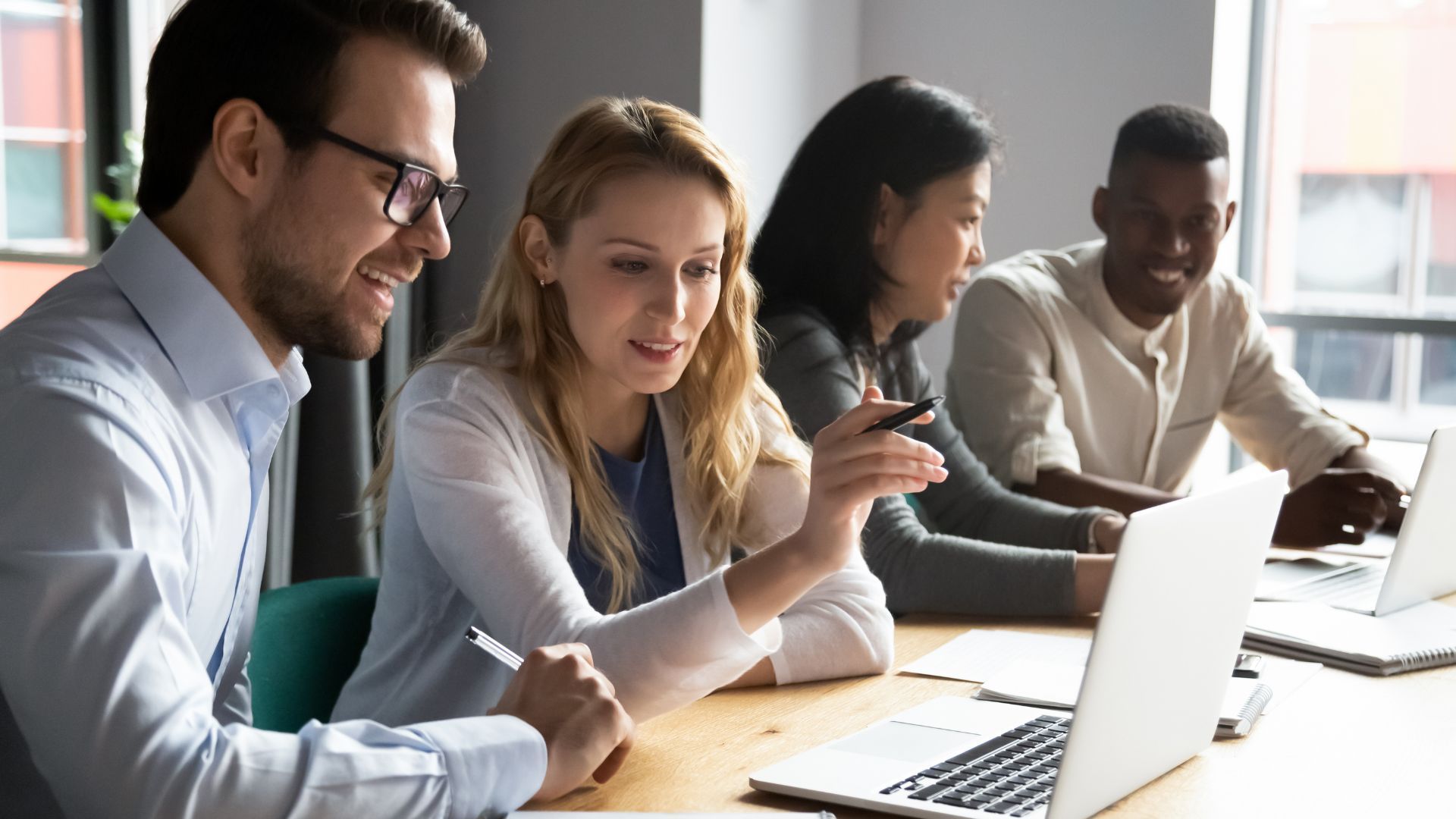 Four people are collaborating at a table, looking at a laptop screen. The woman points to the screen, and others smile.