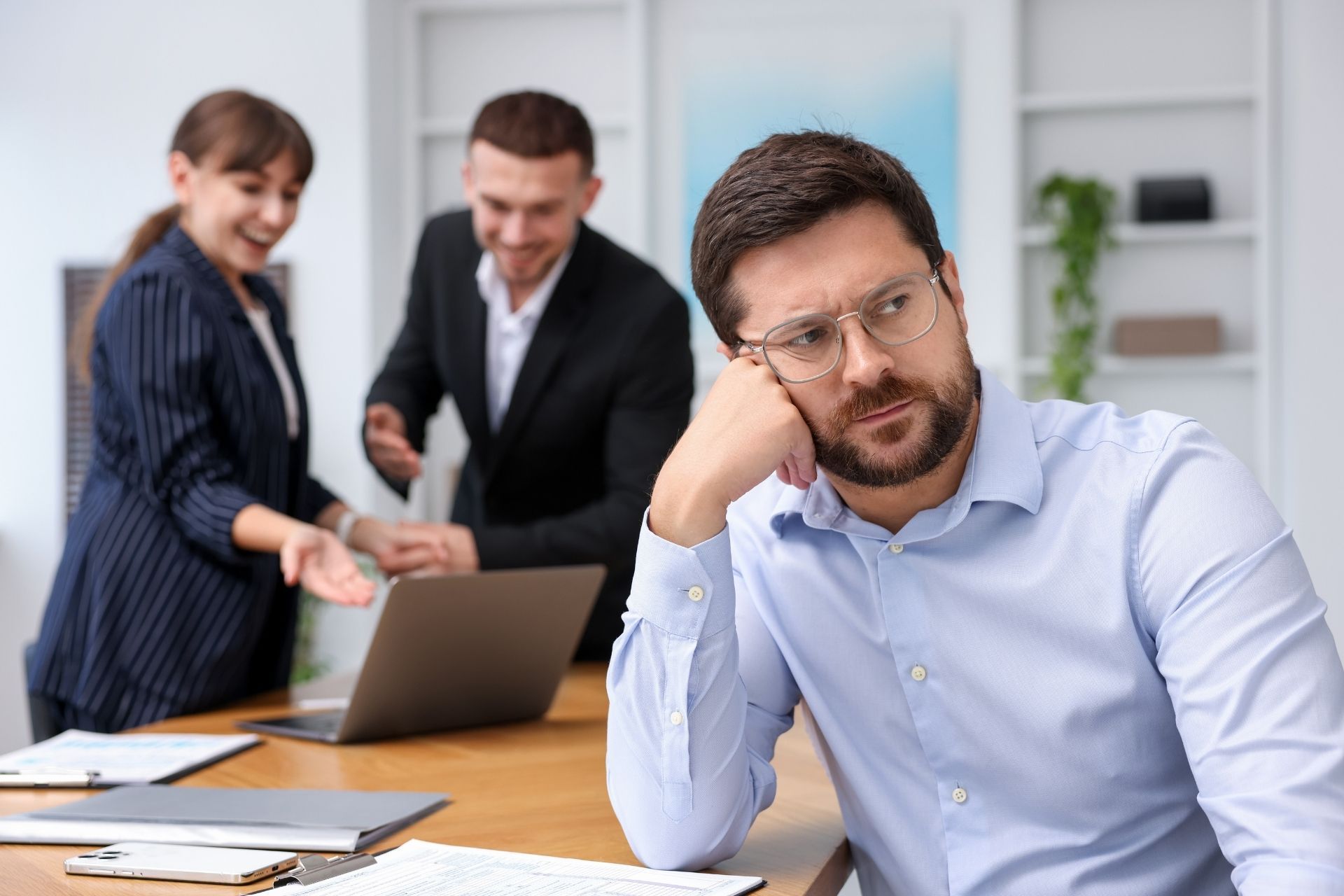 A man sits at a desk looking bored and frustrated, while two colleagues behind him talk and smile at a laptop.