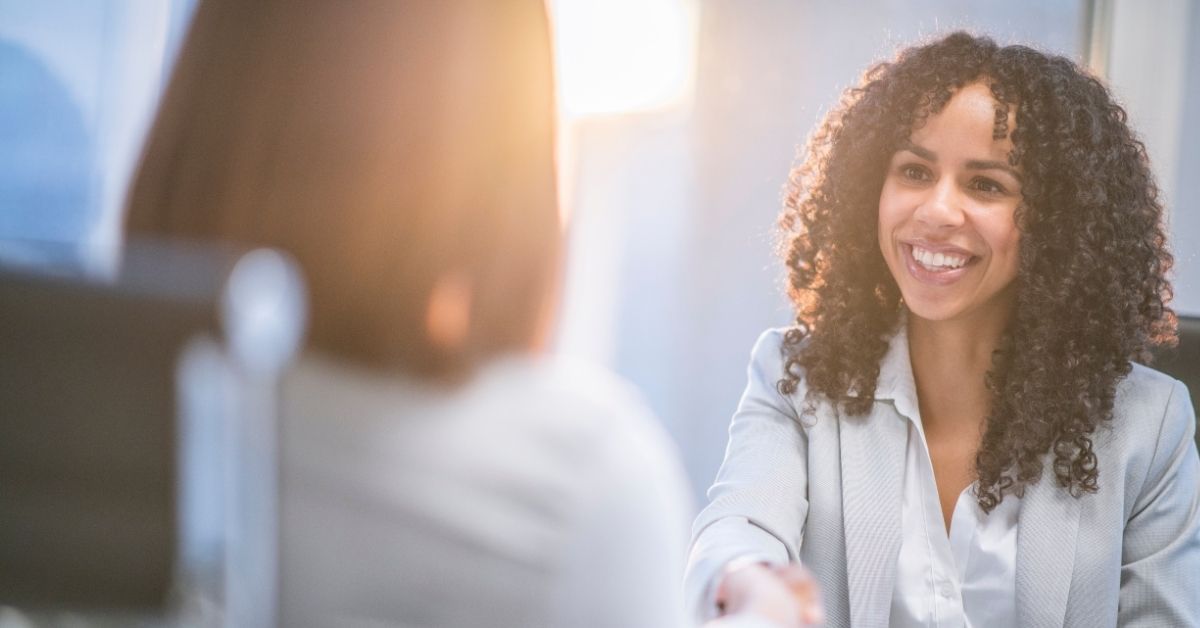 Woman in a light blazer smiling, shaking hands with someone out of frame, in a well-lit office.