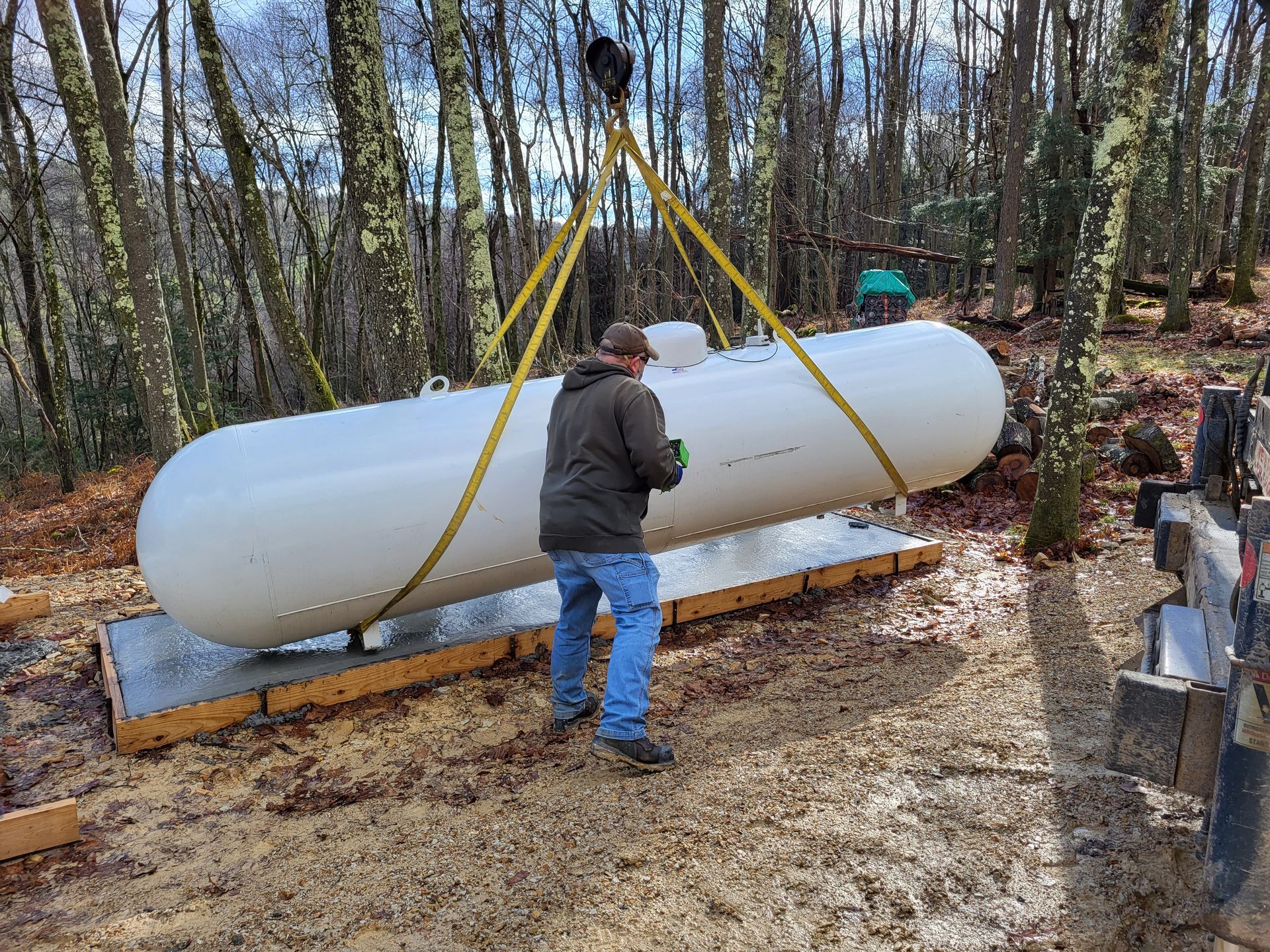 A person guides a propane tank suspended by crane straps. Tank is white, outdoors in a wooded area.