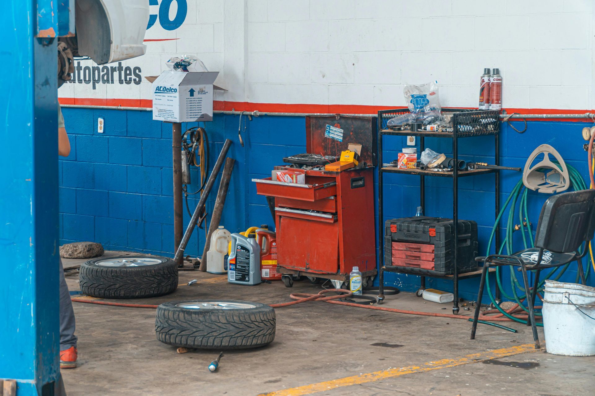 Auto repair shop interior: tires on the floor, red tool chest, blue and red walls, a lifted car, and work tools.