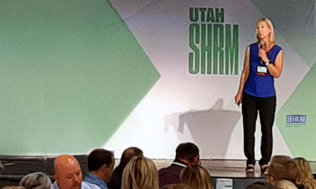 A woman is standing on a stage with a microphone in front of a sign that says utah shrm.