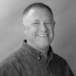 A man in a denim shirt is smiling in a black and white photo.