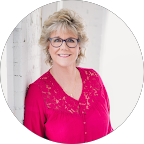 A woman wearing glasses and a pink shirt is leaning against a wall.