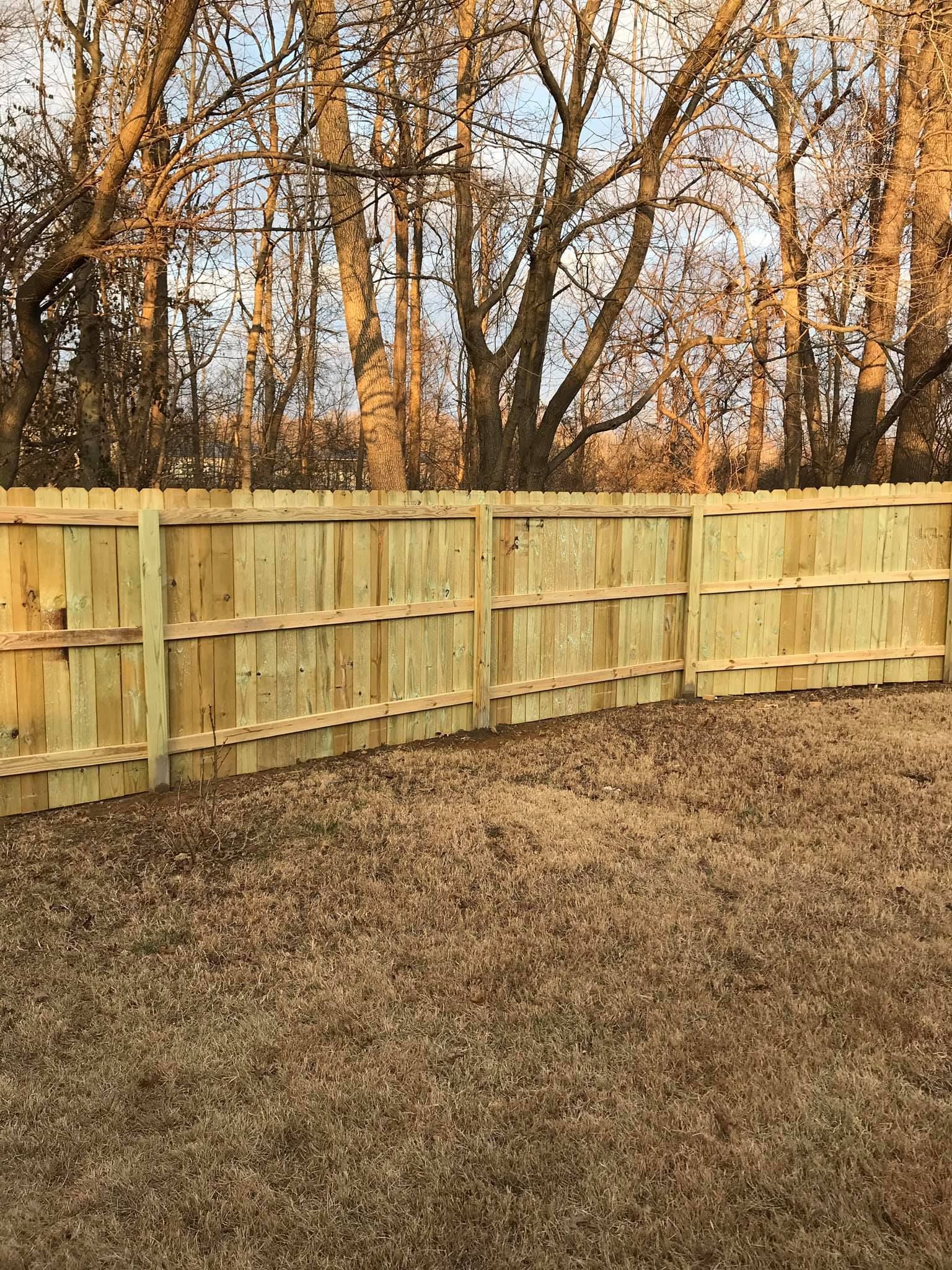 Wooden fence in backyard with brown grass and trees in the background.