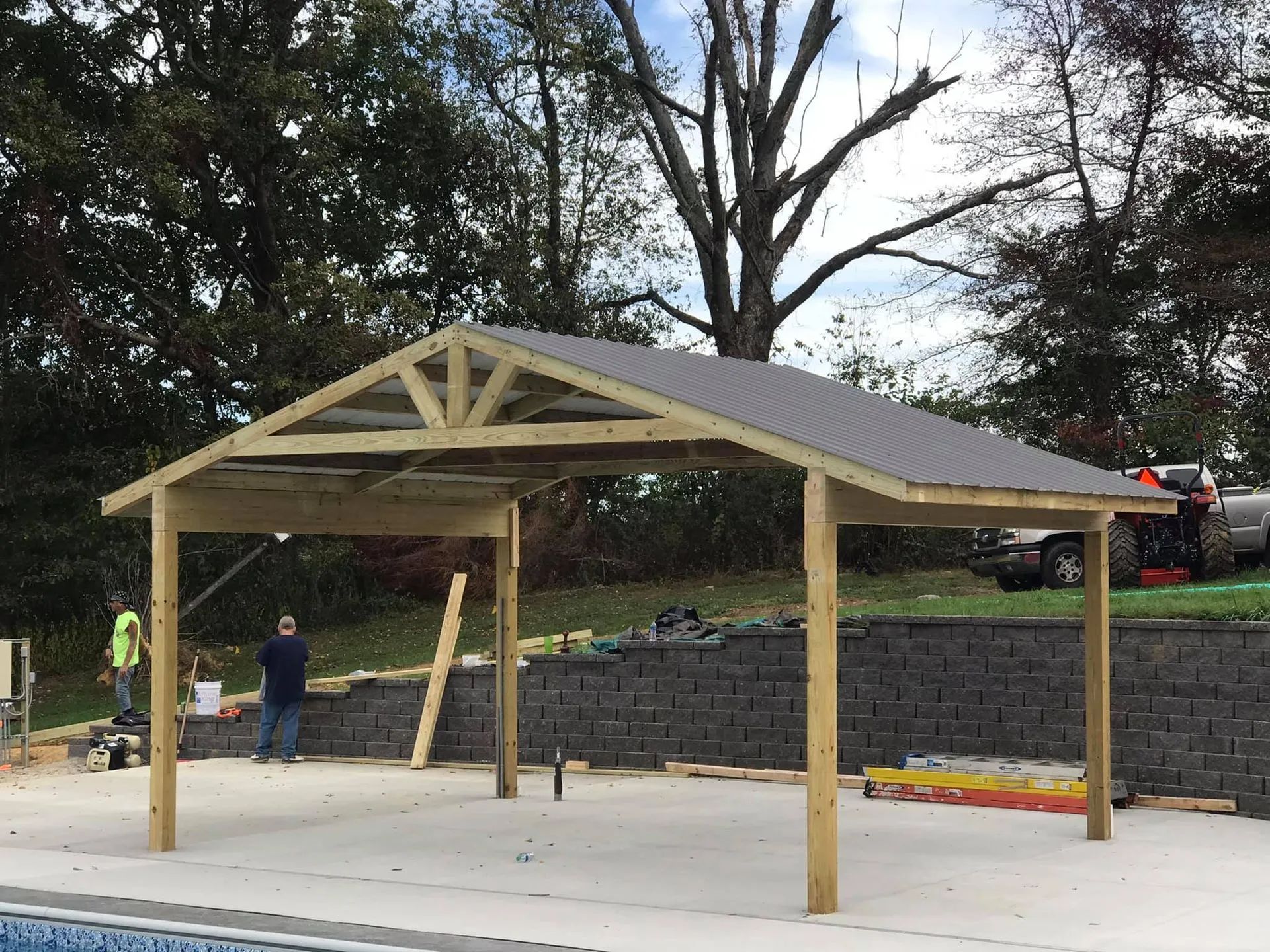 Wooden poolside canopy under construction; workers, gray roof, brick wall, trees.