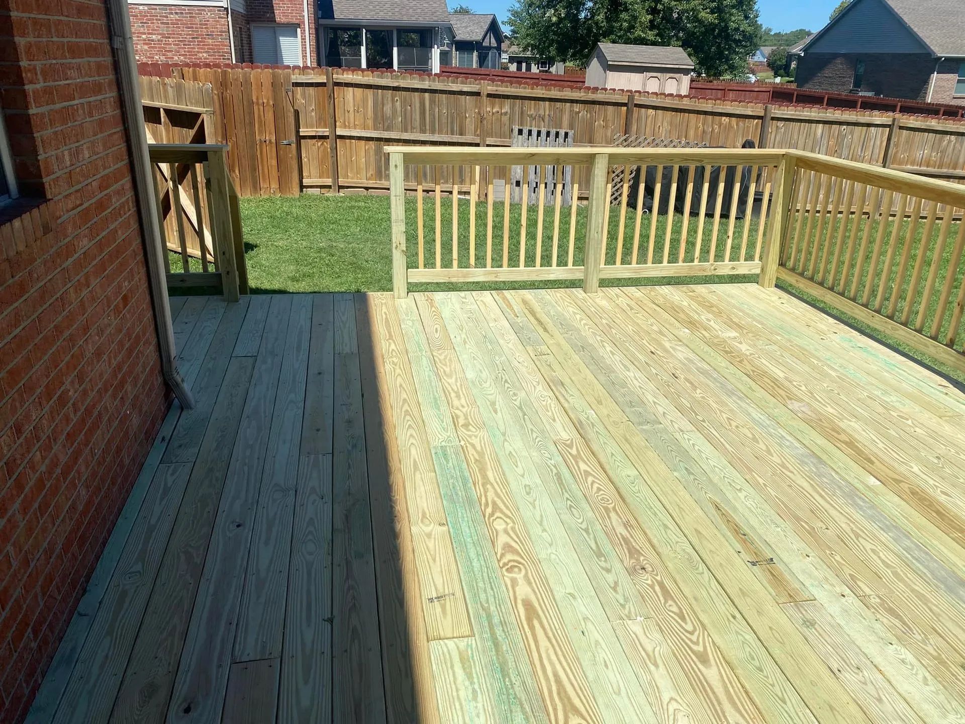 Wooden deck with railing, connected to a brick building next to a fenced yard.