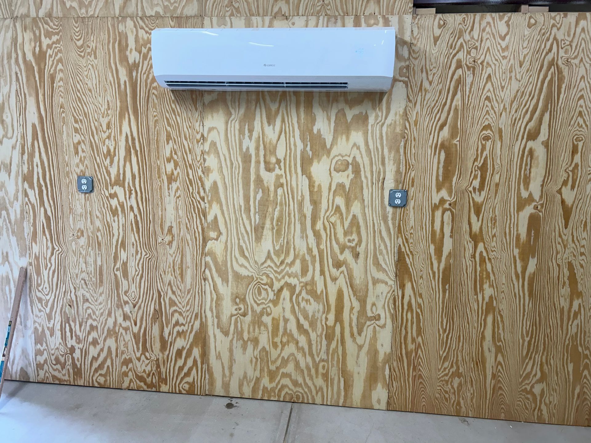 White air conditioner mounted on plywood wall with two electrical outlets.