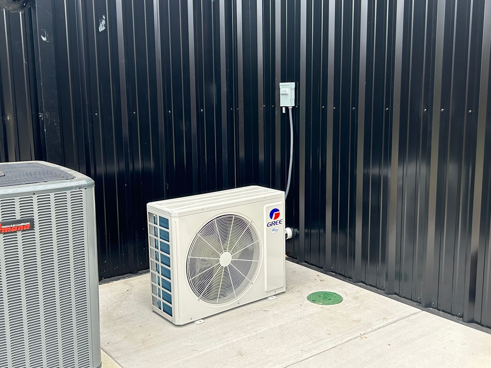Outdoor air conditioning units near a black metal wall. Electrical outlet is visible.