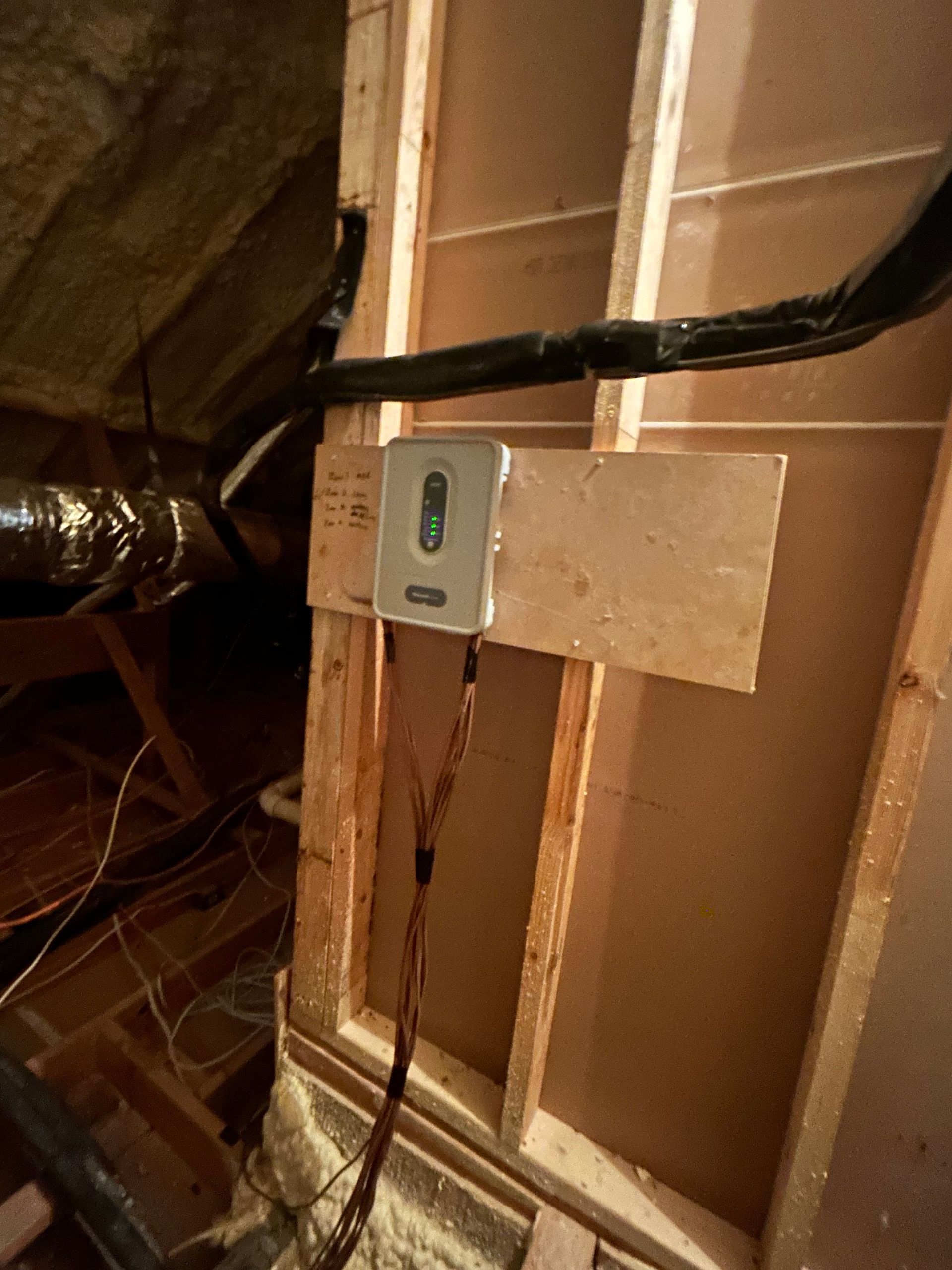 Gray box attached to a wooden board on a wall. Black cables and ductwork in the background.