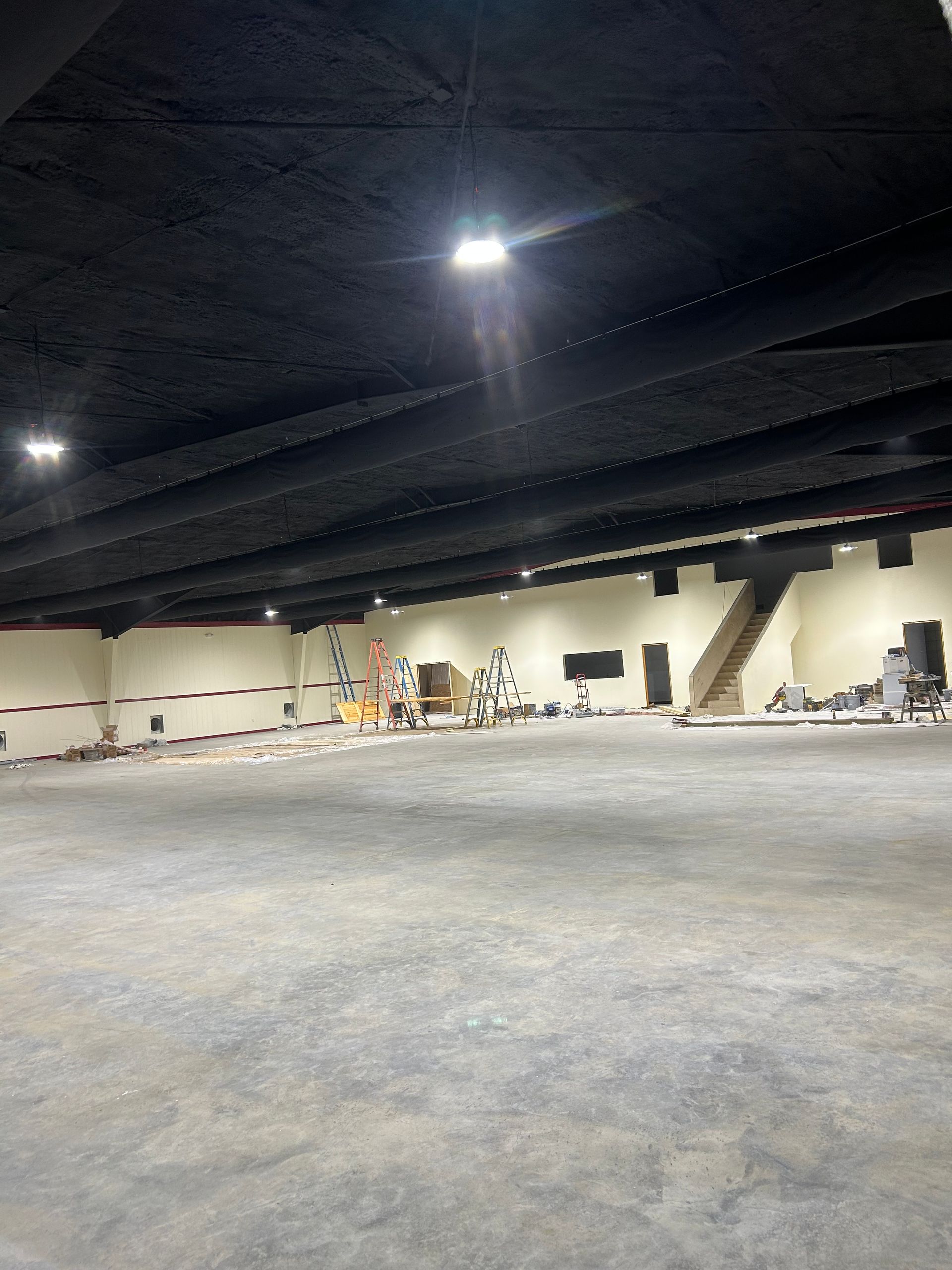 Empty, large room with black ceiling, beige walls, and concrete floor. Stairs and equipment visible.
