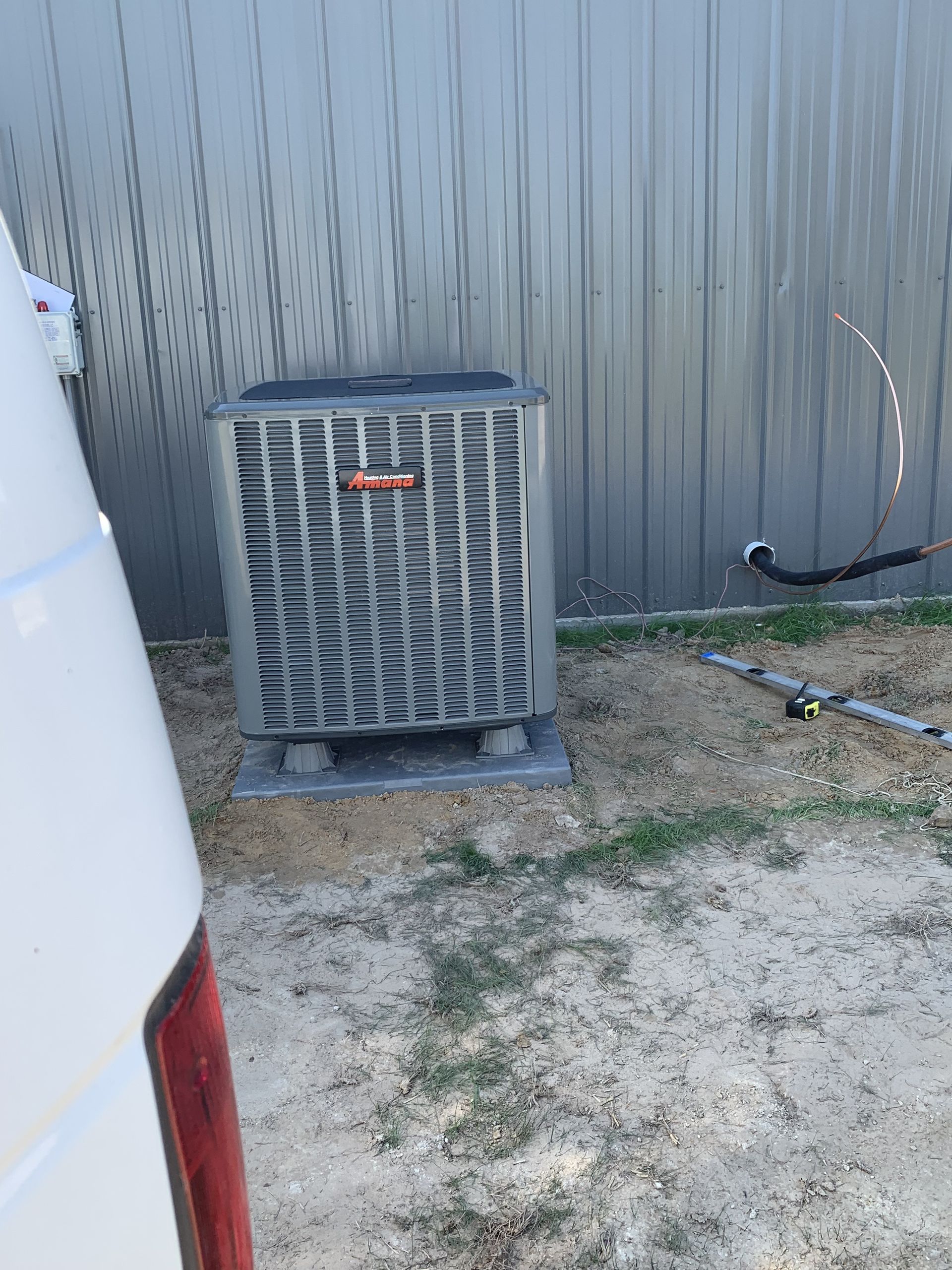 A Goodman air conditioning unit on a concrete pad next to a corrugated metal fence.