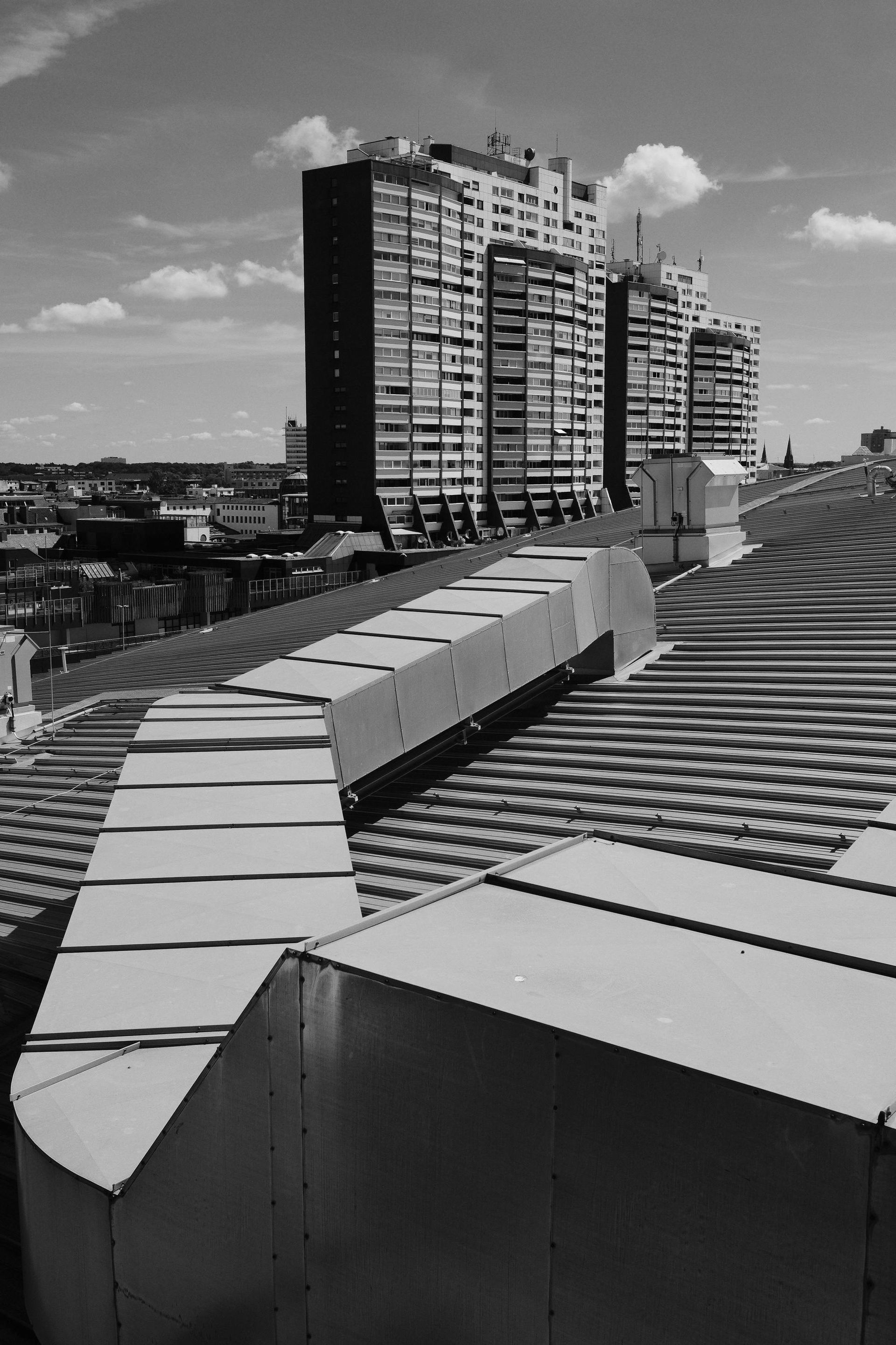 Rooftop view of metal ventilation ducts and tall apartment buildings under a partly cloudy sky.