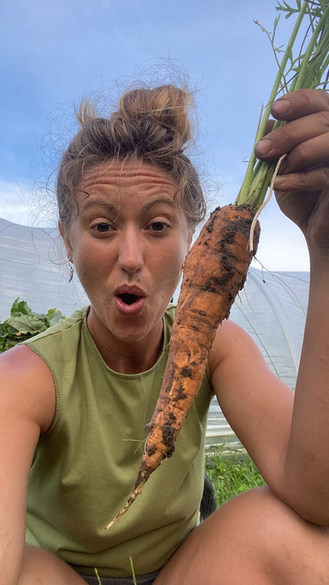 Woman holding large carrot in a garden, surprised expression.