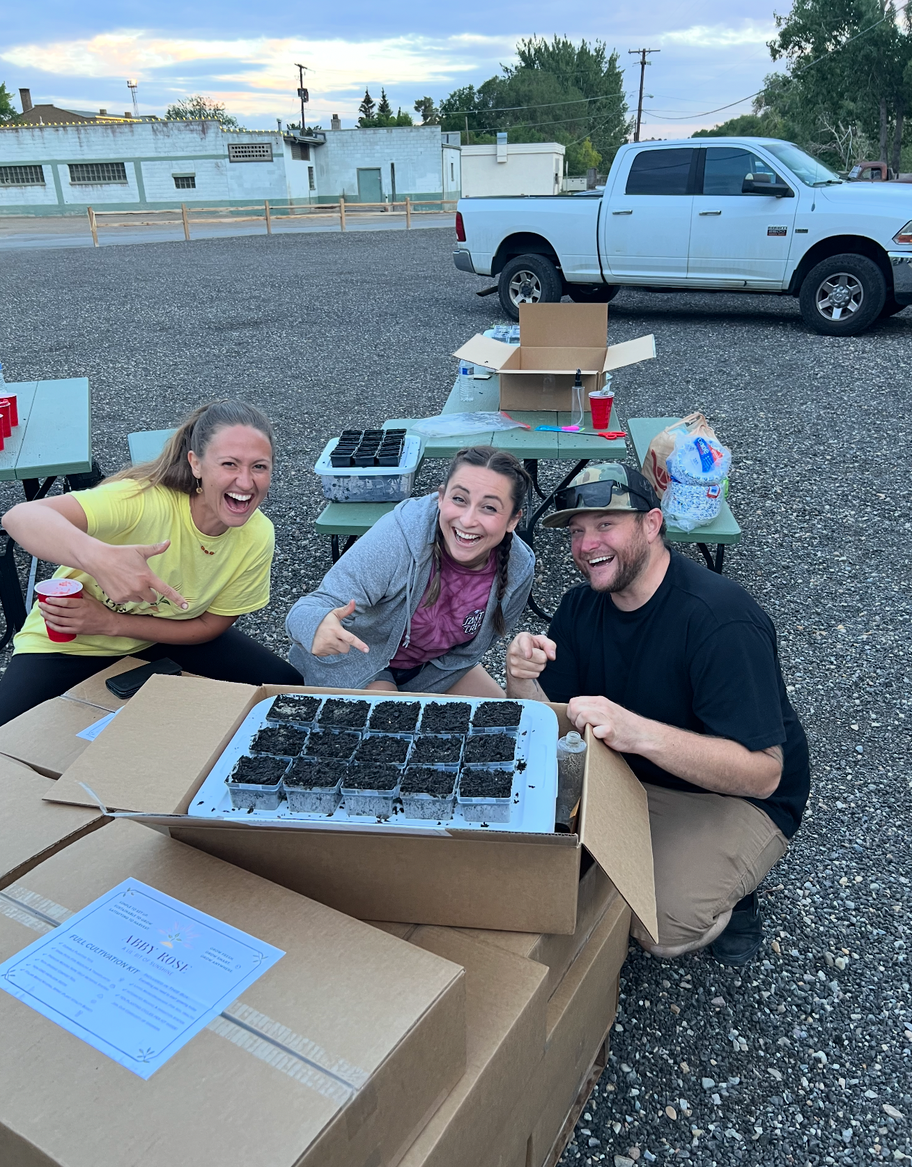 Three smiling people showing off a tray of seedlings outdoors by boxes and a truck.
