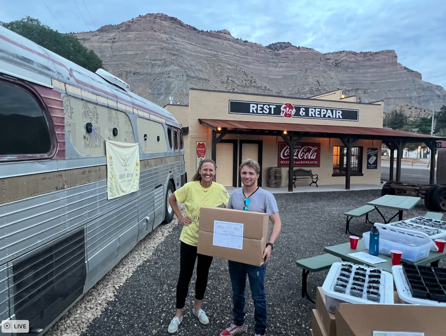 Two people holding boxes in front of a repair shop and bus; mountainous backdrop.