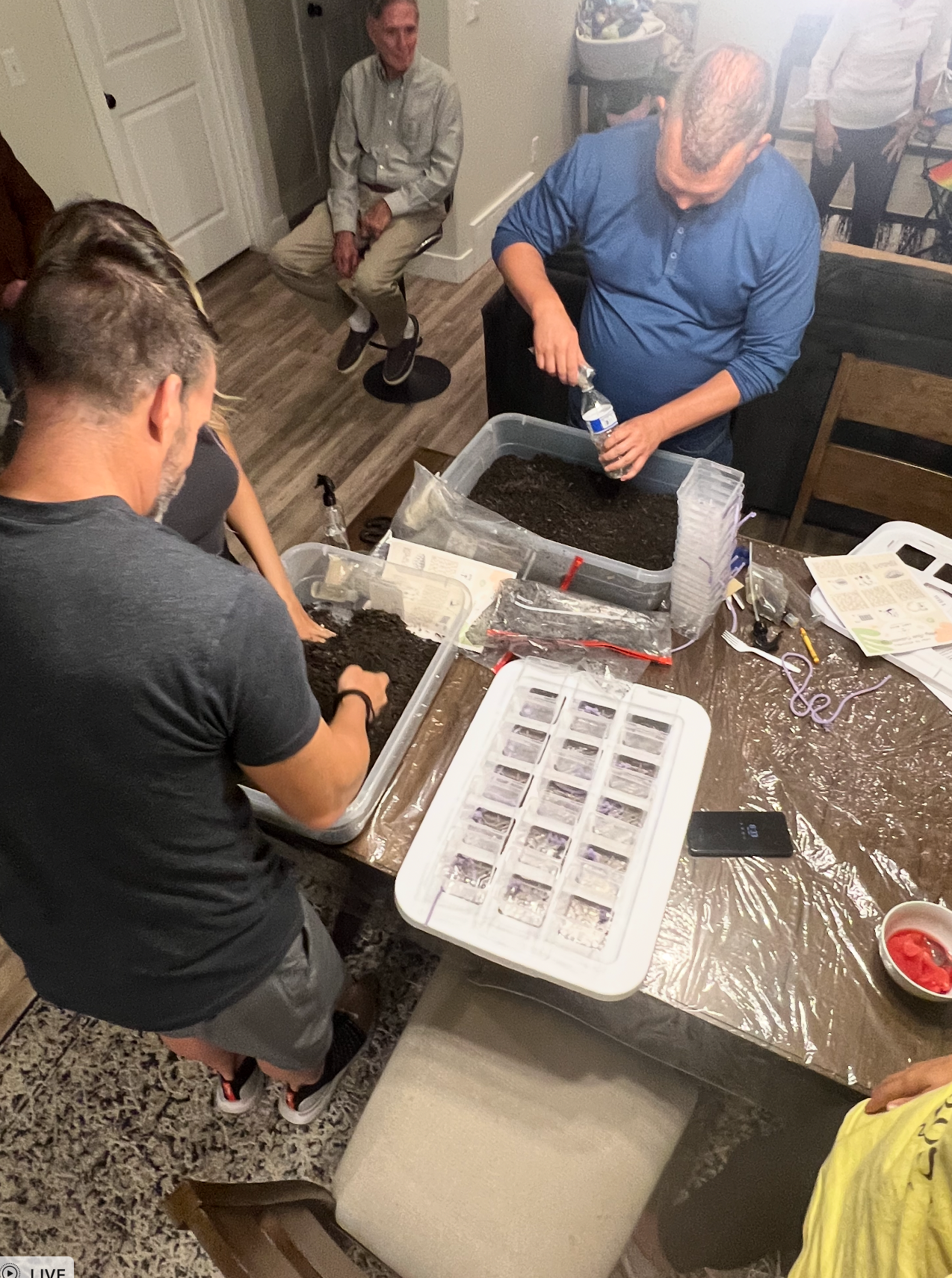 People planting seeds indoors at a table. Men and women fill trays with soil.