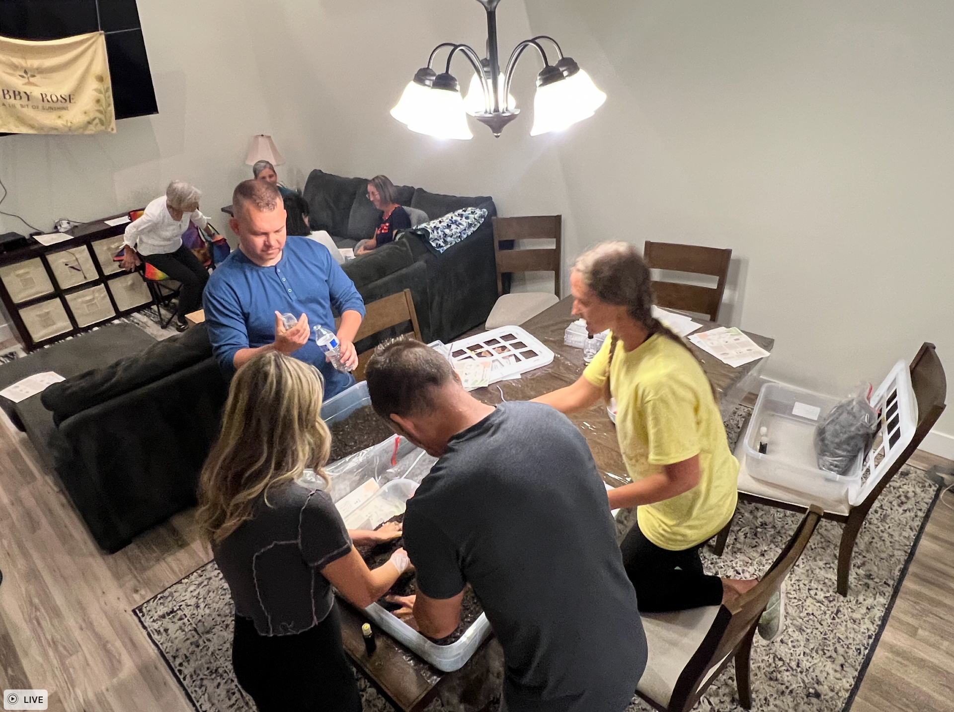 People sorting items around a table, living room setting, others seated on a couch.