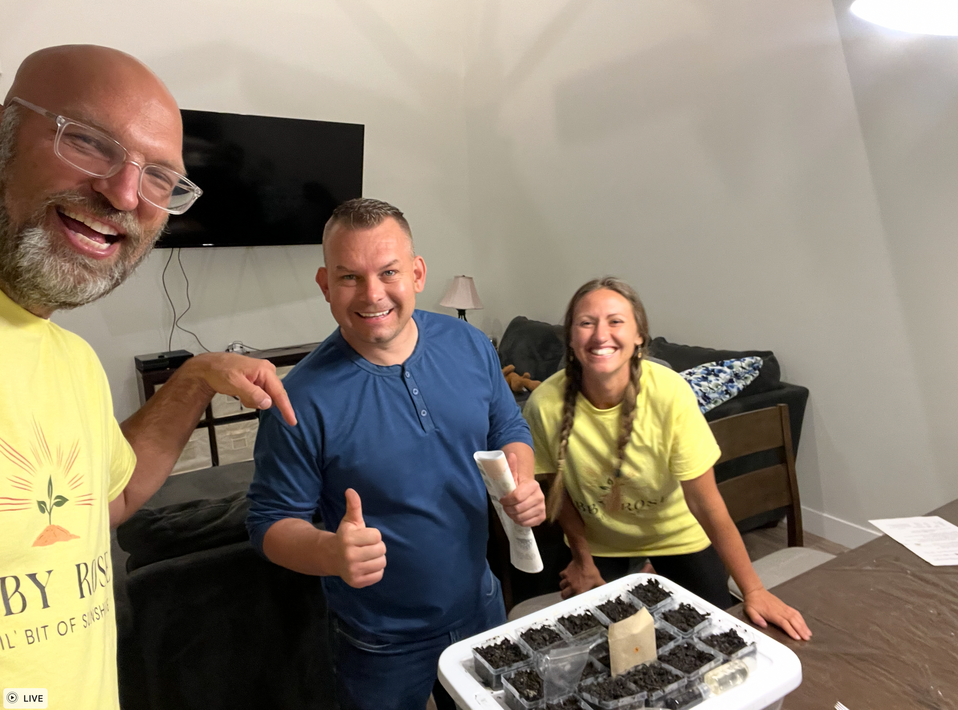 Three people smiling around a container of plants indoors. One man points, another gives a thumbs up.