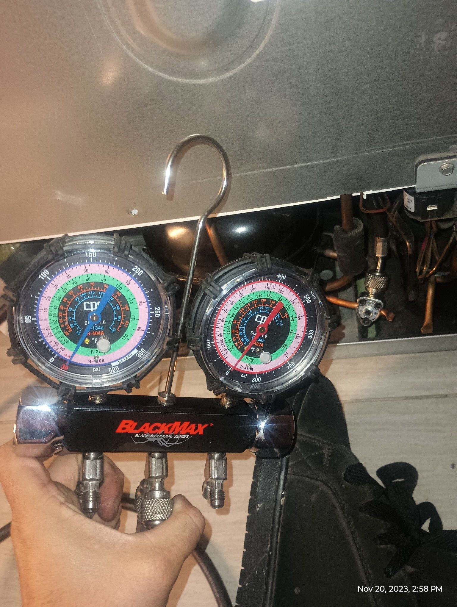 Blue-and-red gauge manifold held in front of an appliance, with a hook above and a hand gripping it