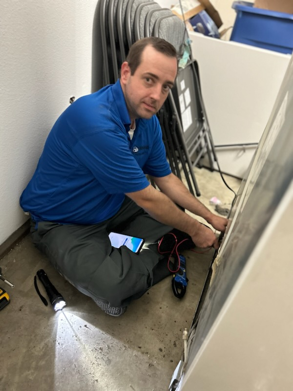 Technician kneeling on a floor, working on cables beside stacked folding tables in a storage room.