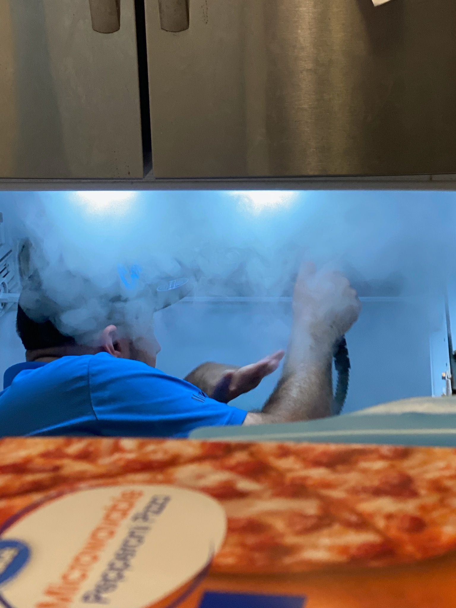 Chef in blue shirt cooking amid steam behind a pizza counter with a pepperoni pizza in the foreground