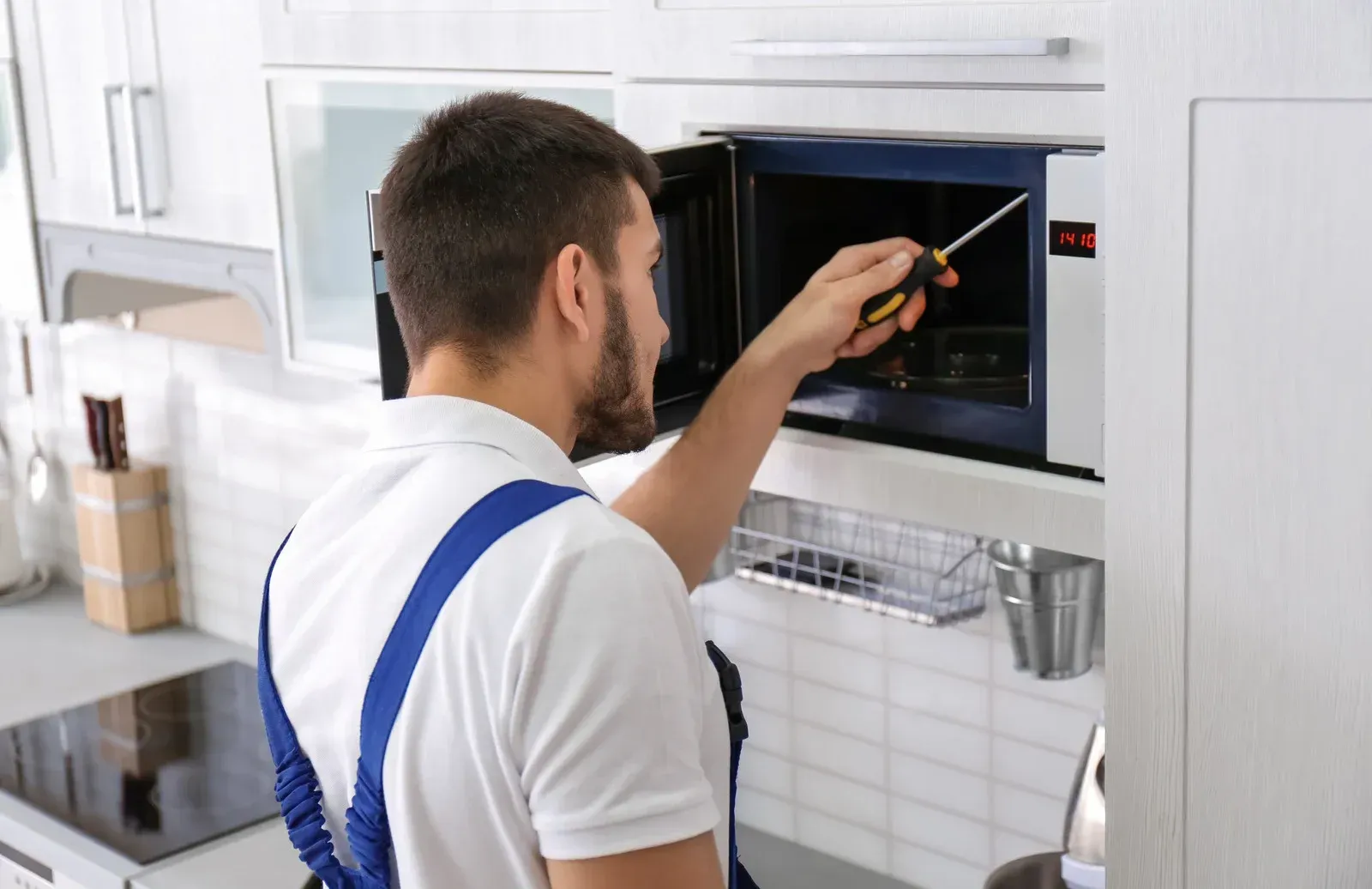 Technician using a screwdriver to repair a built-in microwave in a white kitchen