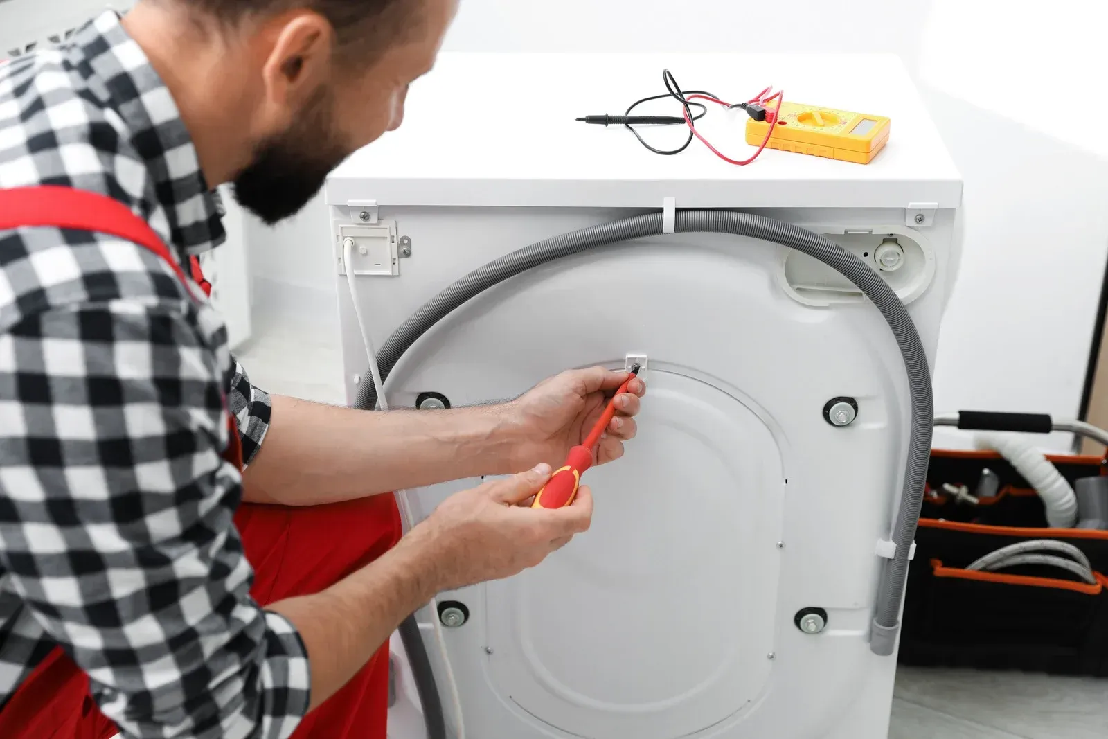 Man repairing a washing machine with a screwdriver, with the back panel open and hoses exposed