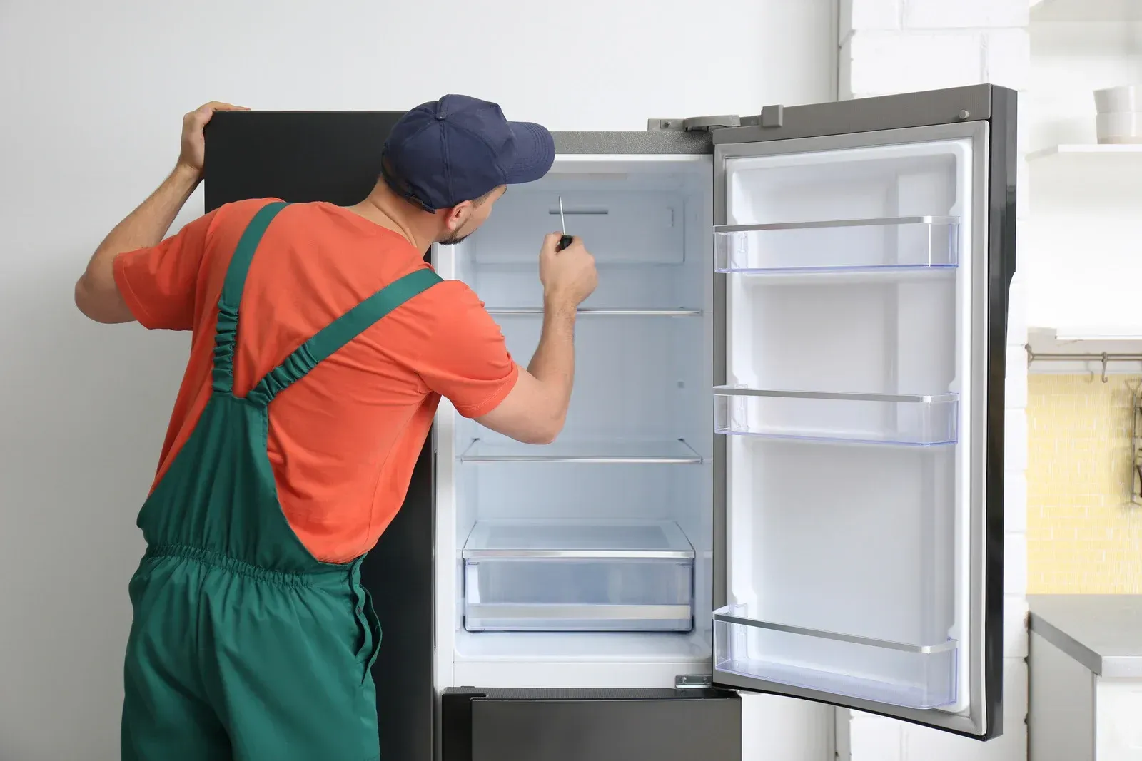 Worker in orange shirt and green overalls inspecting an open refrigerator in a kitchen.