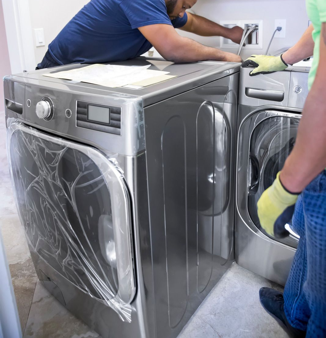 Two workers in gloves move a wrapped front-loading washer and dryer in a room.