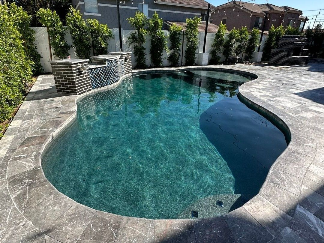 A backyard swimming pool with dark blue water and a stone waterfall feature, surrounded by gray stone pavers and hedges.