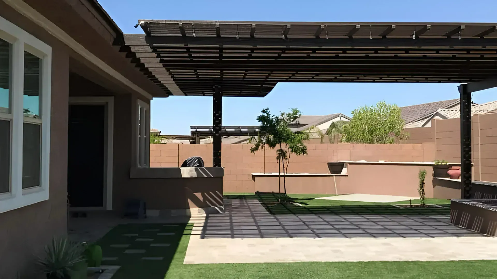 A sunlit patio features a pergola covering a paved area, artificial turf, and a brown wall in a suburban backyard.