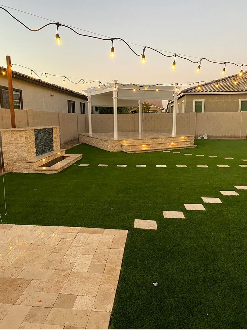 Backyard at dusk with a stone fountain, artificial turf, stepping stones, a raised patio with a pergola, and string lights.