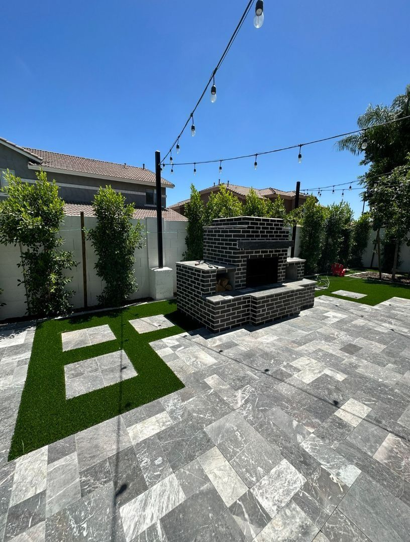 A patio with gray stone pavers, patches of artificial grass, string lights, and a brick outdoor fireplace against a wall.