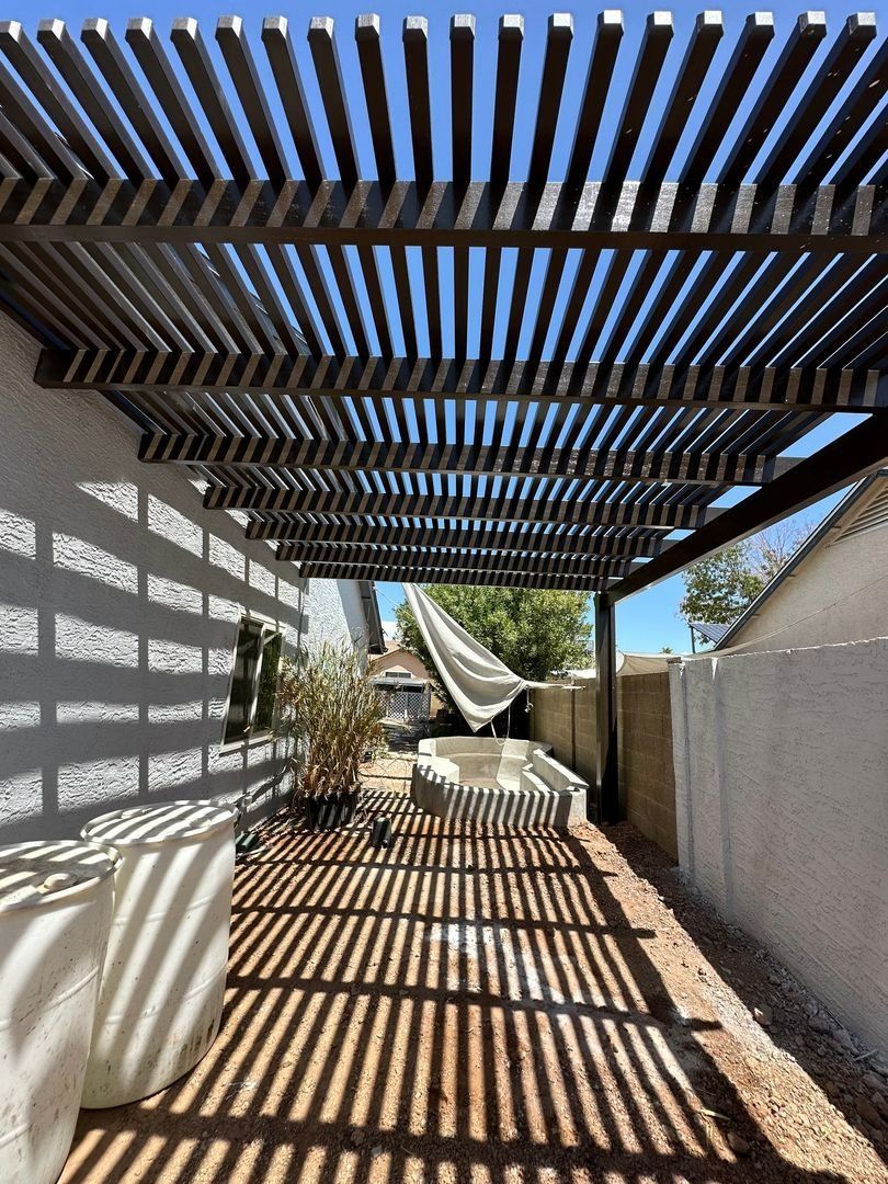 A patio area with a slatted wooden pergola casting striped shadows over a dirt ground, near a stucco wall and small plant.