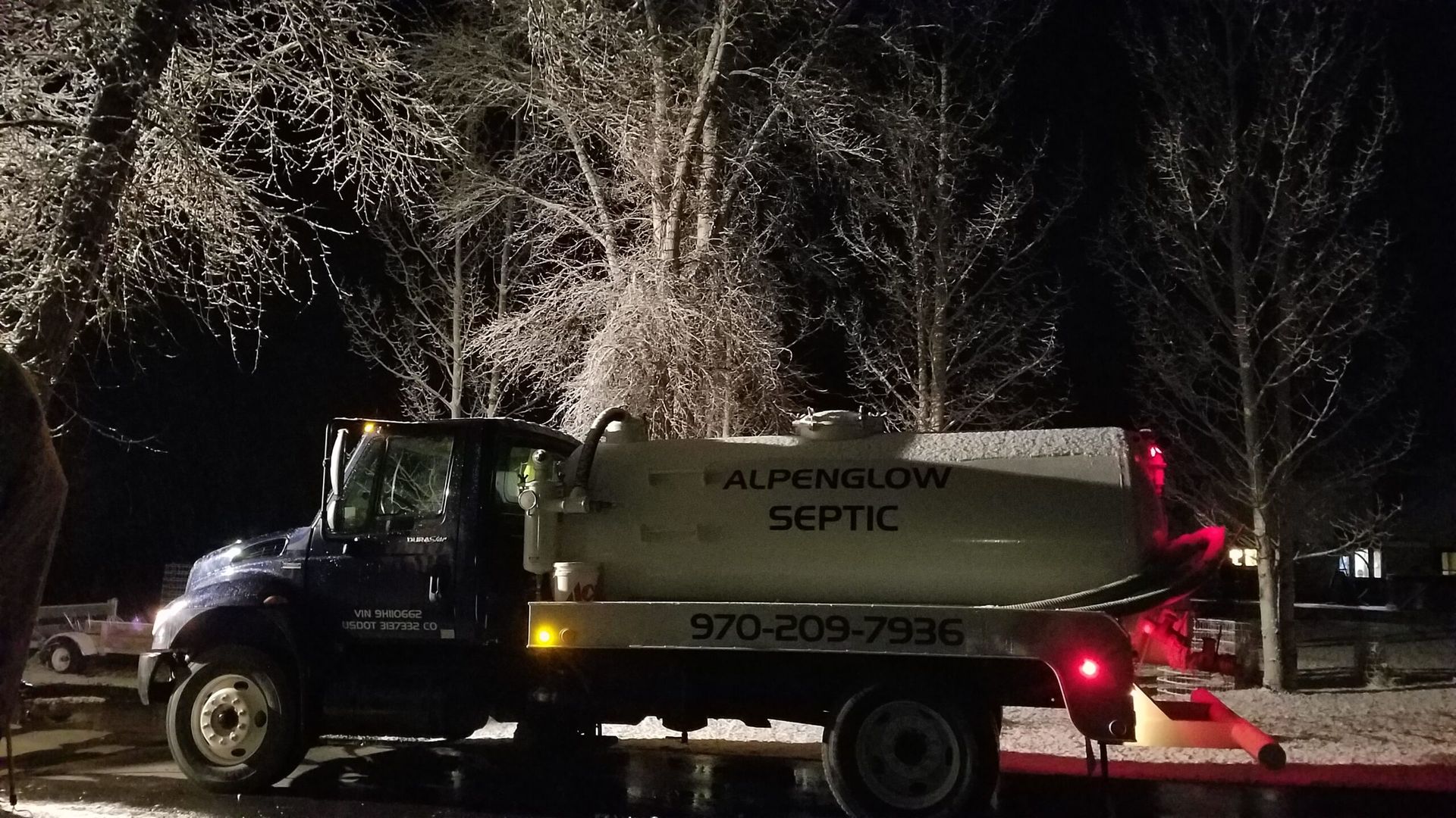 PumpEnging Septic truck parked at night under trees, with rear lights glowing and emergency hazard lights on.