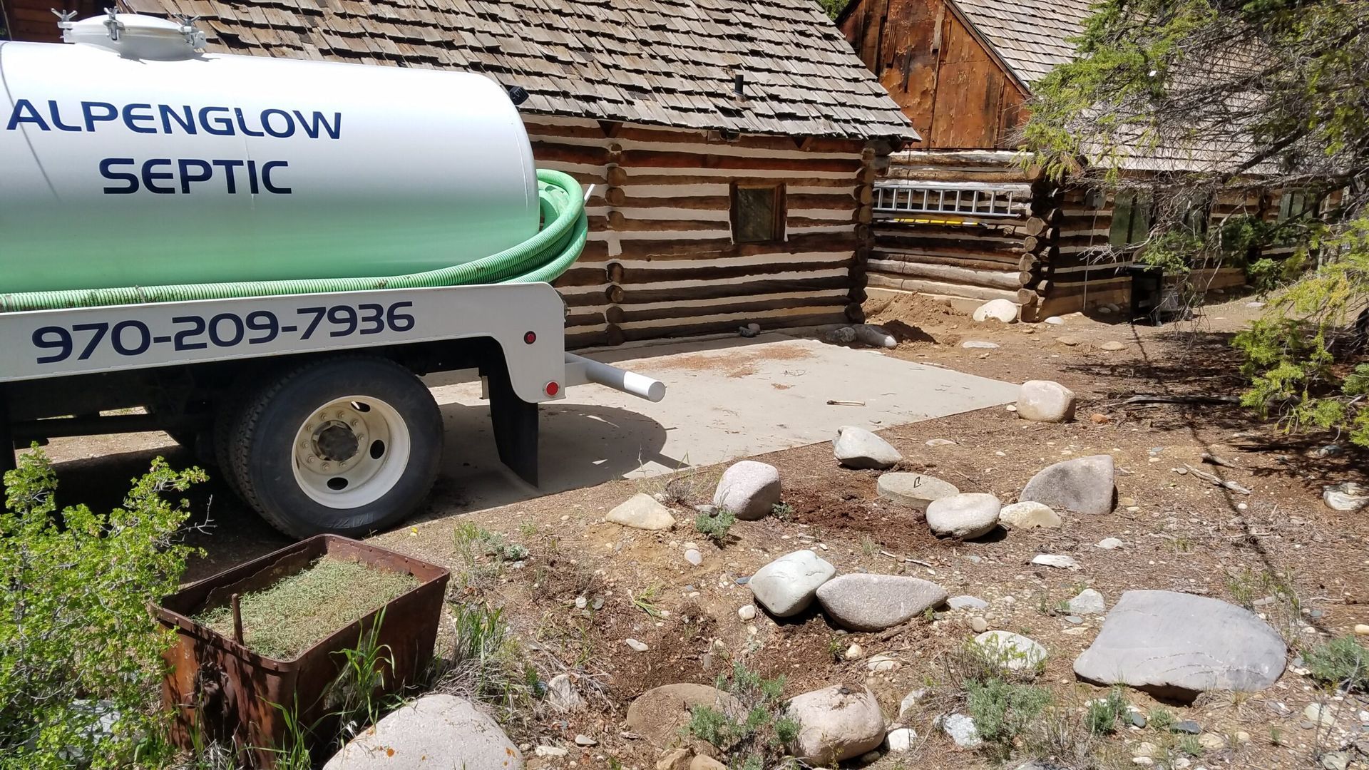 Septic tanker truck beside a rustic cabin and stone path in a wooded yard