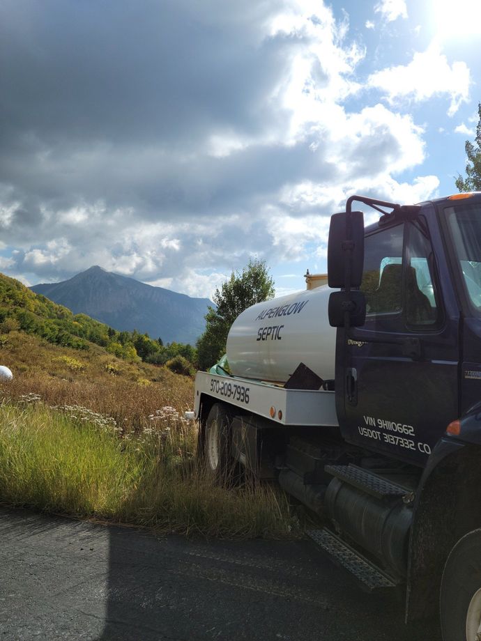 White work truck parked on a roadside beside tall grass, with mountains and cloudy sky in the background