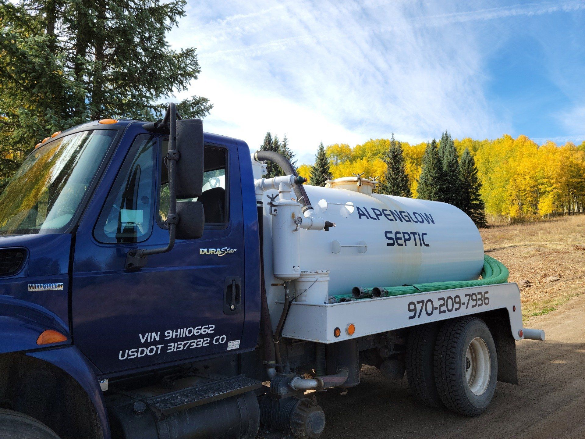 Blue septic truck parked in a field with autumn trees in the background