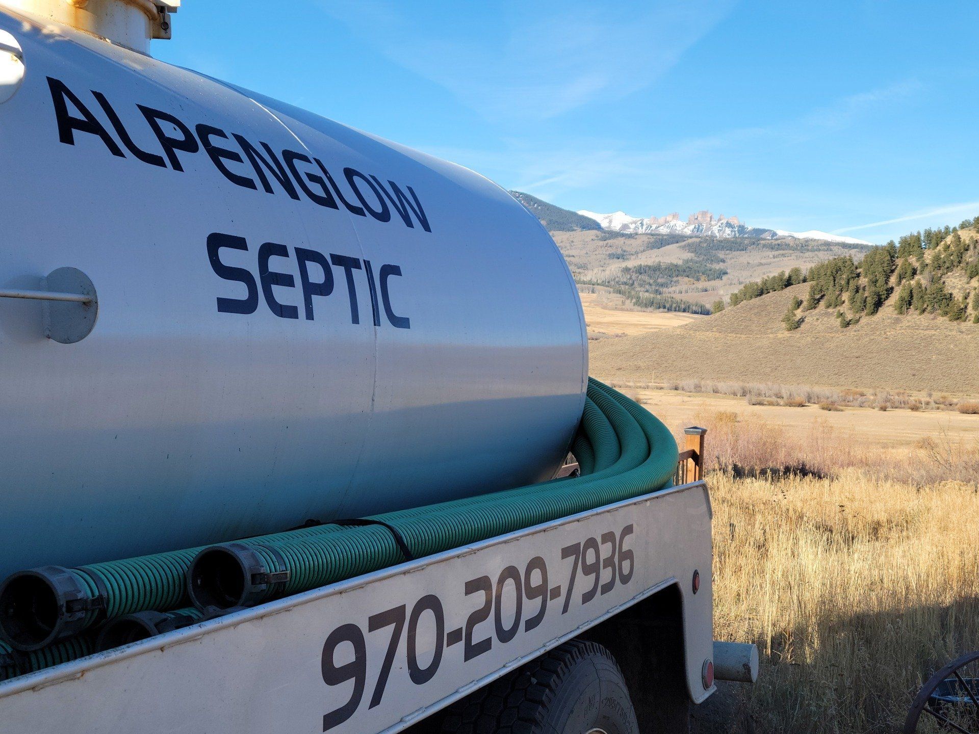 White septic truck tank with “ALPENGLOW SEPTIC” and phone number, parked by a grassy mountain field.