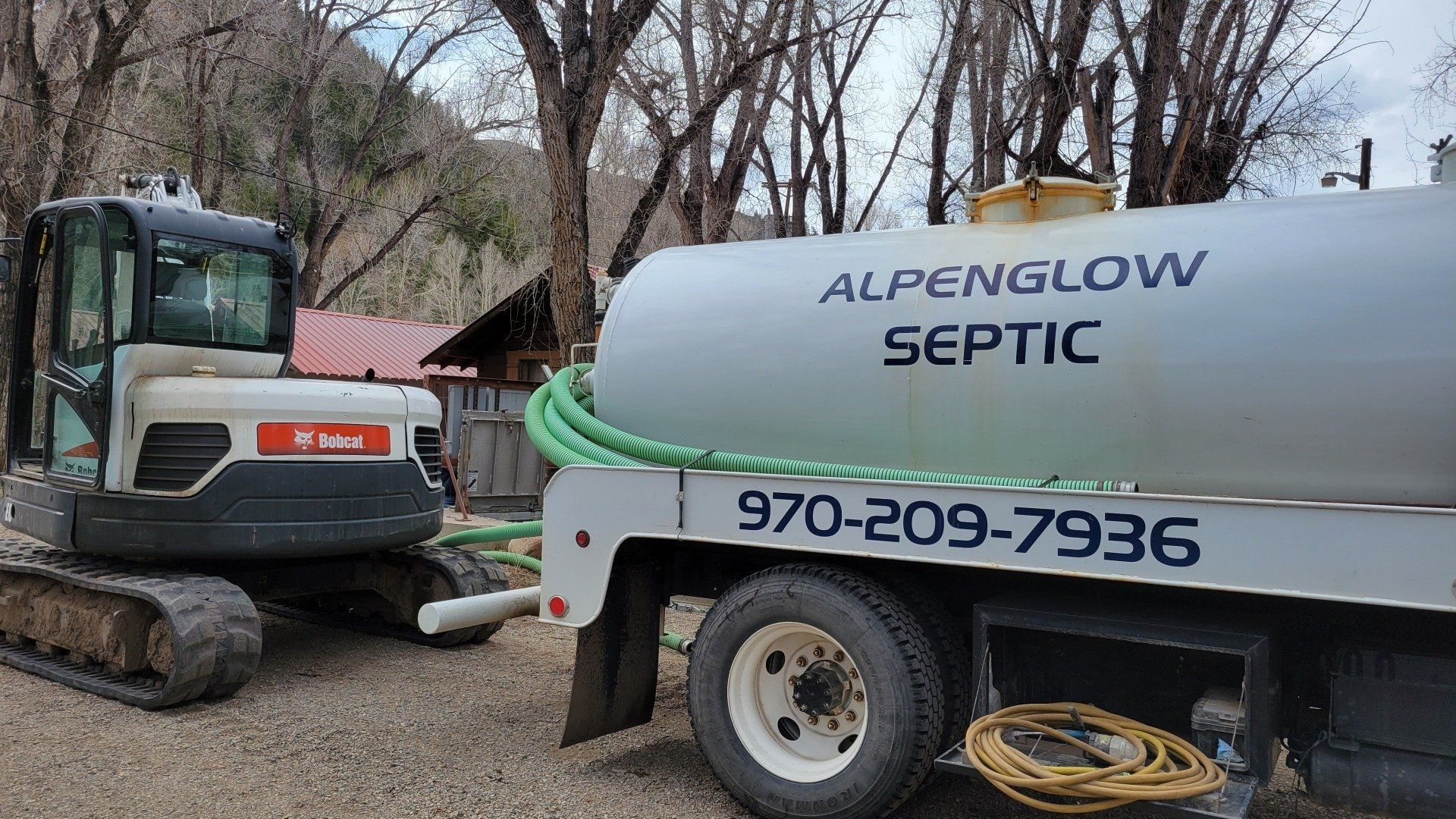 Excavator beside ALPENGLOW SEPTIC truck with hose and phone number in a wooded area