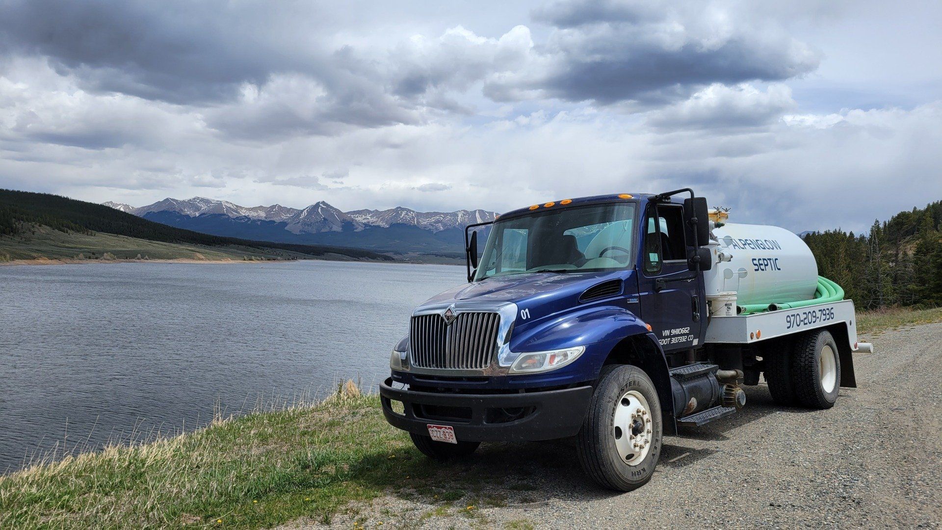 Blue water truck parked on a gravel road beside a lake with mountains and cloudy sky in the background
