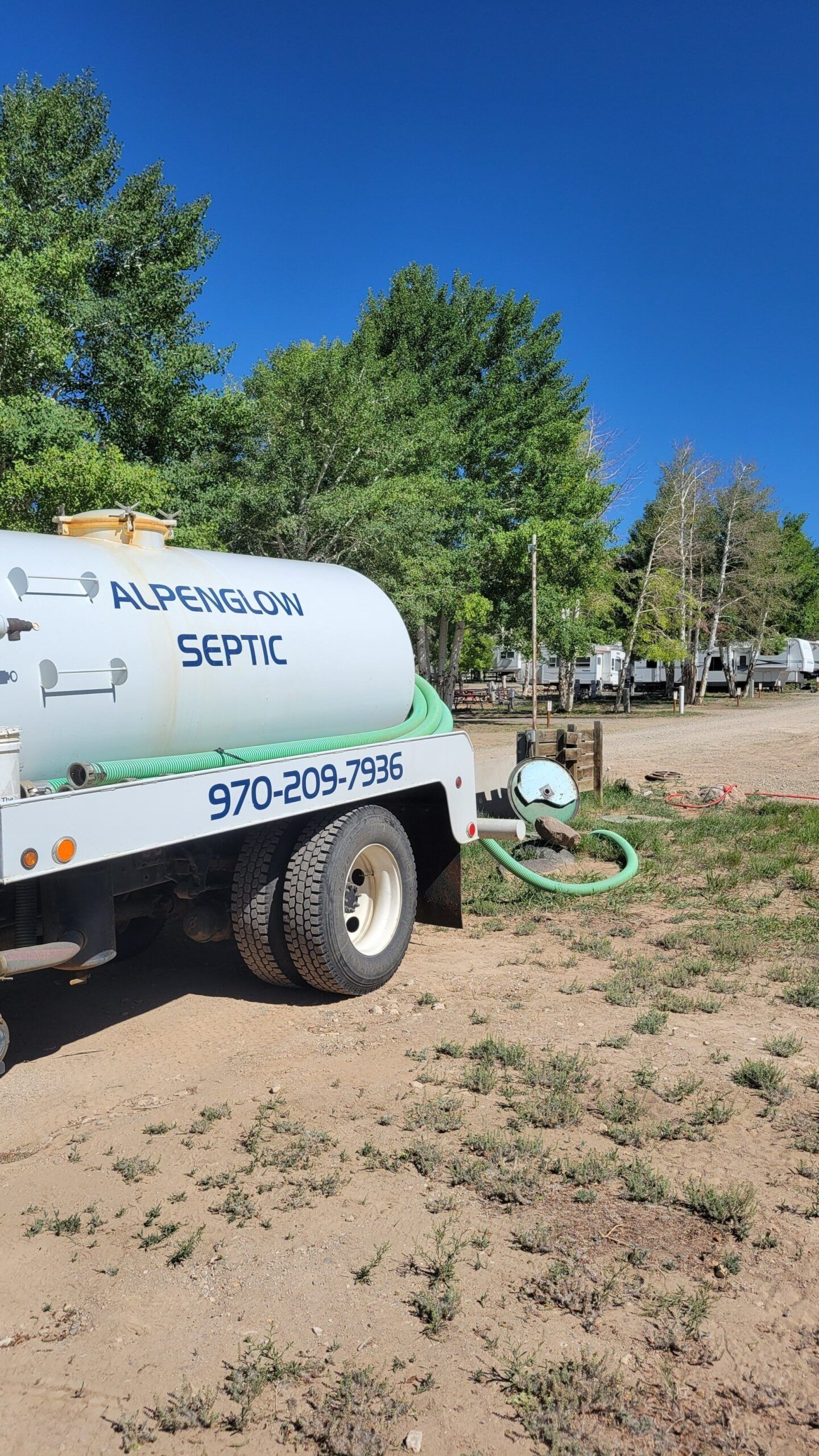 White service truck with green hose in a dry yard under trees and blue sky