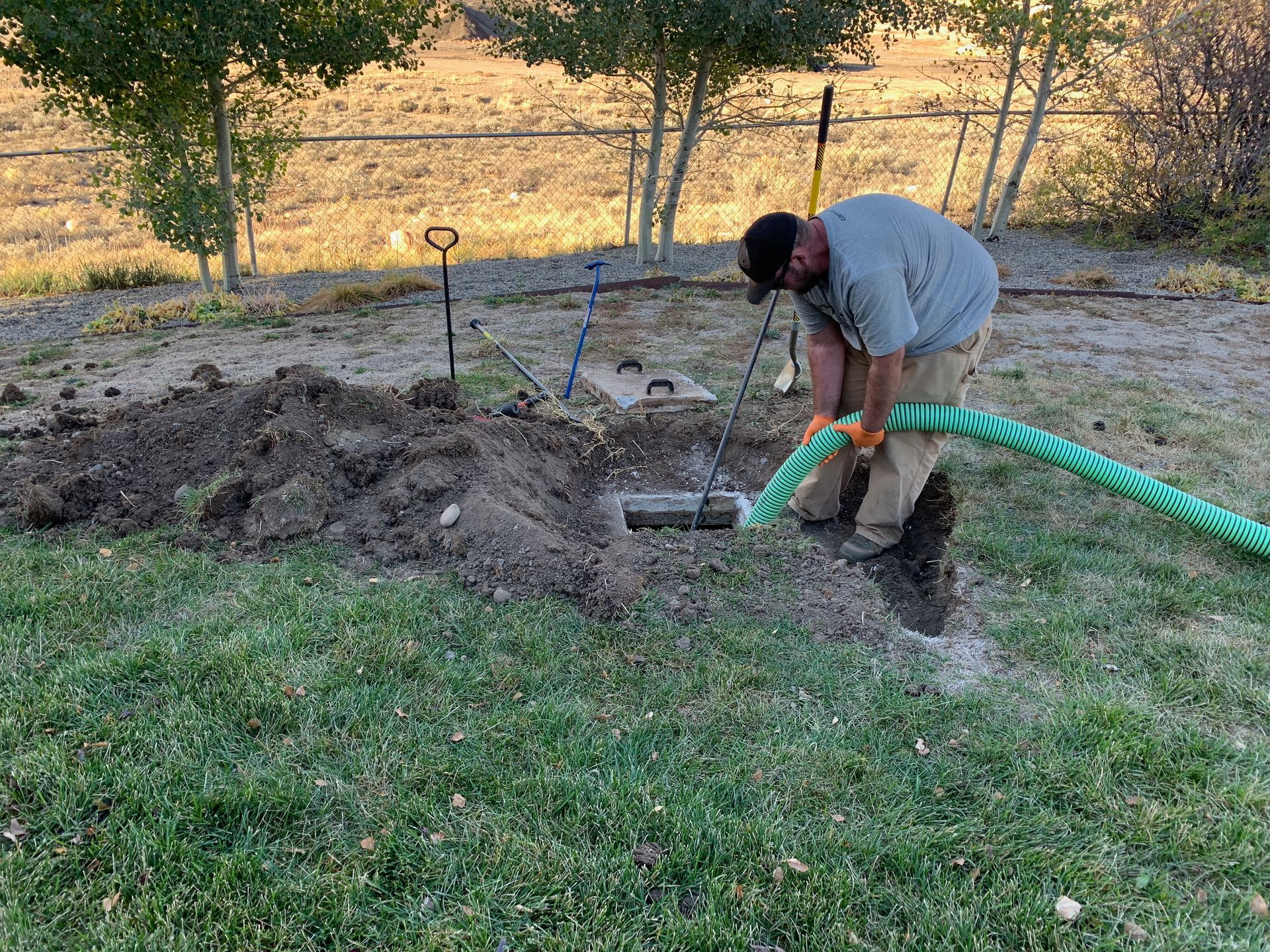 Person digging a trench with a green hose in a grassy yard near trees