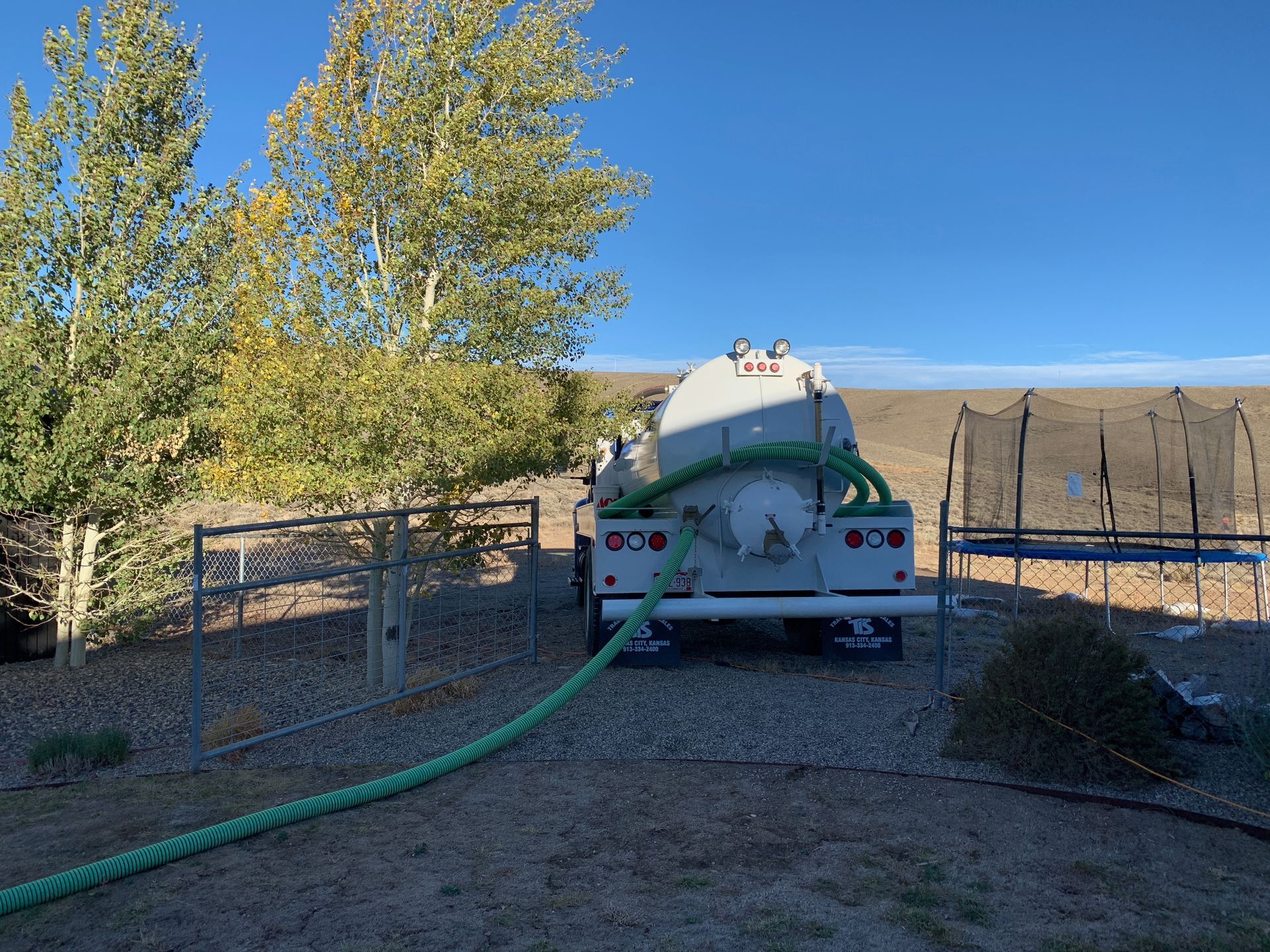 Water tanker truck parked in a rural field, with a green hose and trees nearby under a blue sky.