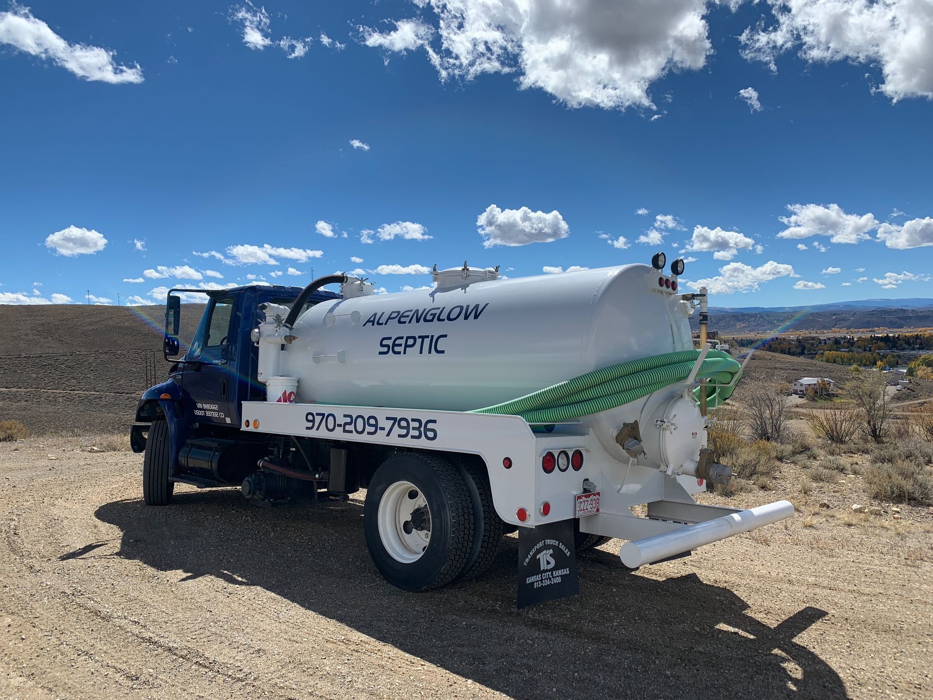 White septic truck on a dirt road under a blue sky with clouds, mountains in the distance.