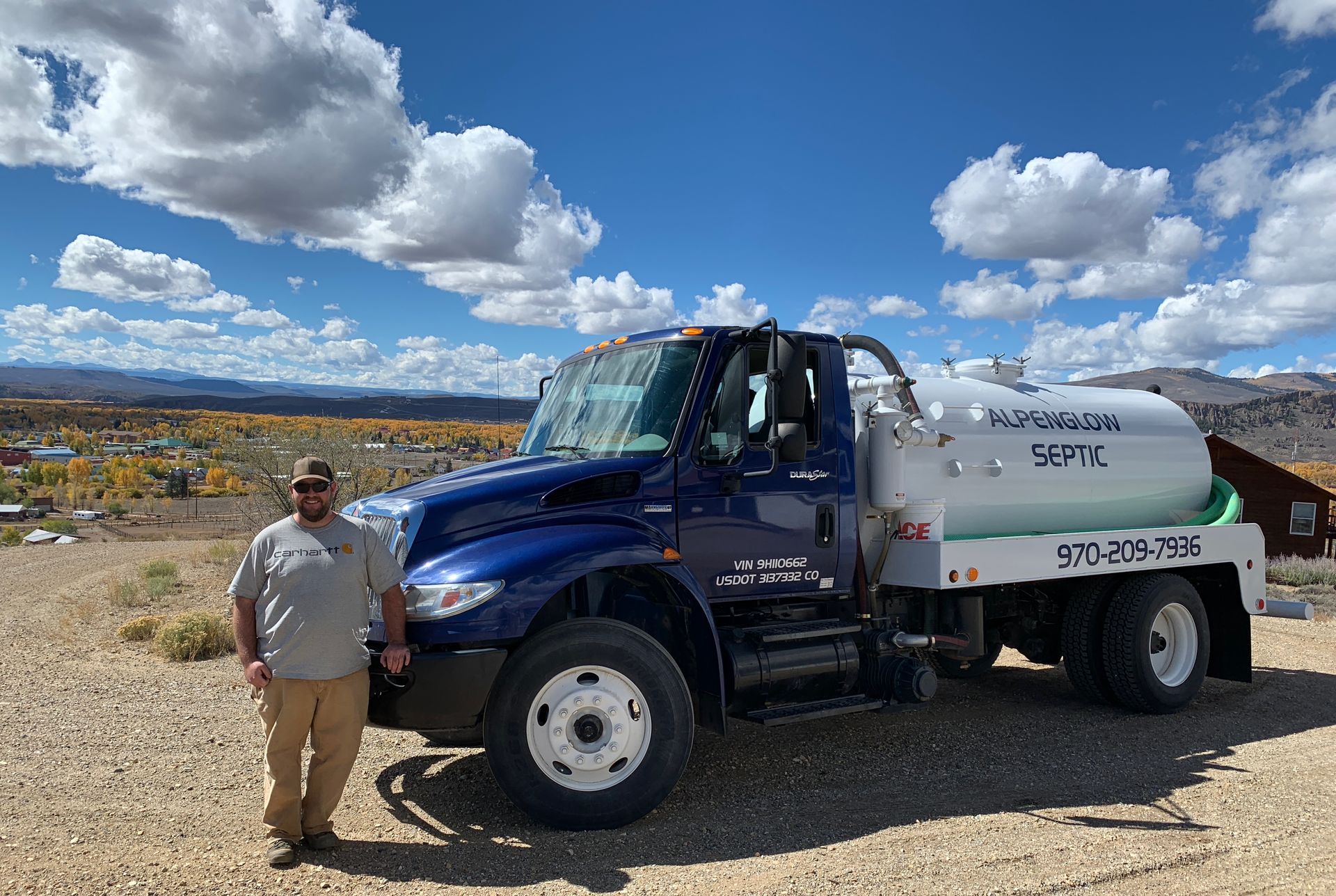 Man standing beside a blue-and-white tanker truck on a dirt road under a cloudy sky